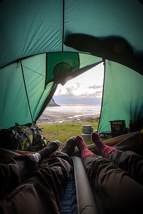 The image is largely dominated by black hues and portrays an outdoor setting. It showcases a person's legs, clothed in red socks, extending from inside a tent. The individual appears to be relaxing on a camping mat with various hiking equipment scattered around them. Nearby, there is a stack of cooking pots and a green backpack with straps. Laying on the grass, there is a book. The tent is pitched with a view of the ocean and the beach, suggesting a camping trip. The sky is visible, indicating a clear day, perfect for outdoor activities. Furthermore, there is a tarpaulin and an unidentified black object with a green background present in the image. The scene gives off a peaceful vibe of a person enjoying their time in nature.