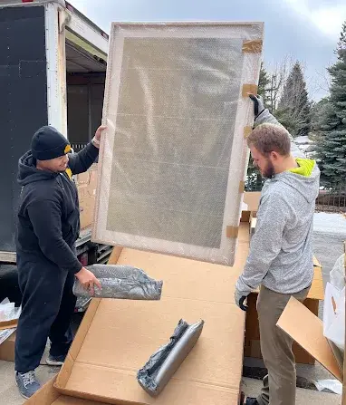 Two men unload a large framed panel from a truck outdoors, surrounded by packing cardboard and wrapped parts.