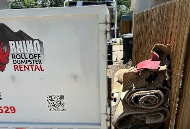 White Rhino Roll Off Dumpster Rental truck beside a fence, with rolled-up carpets and debris piled in a narrow alley.