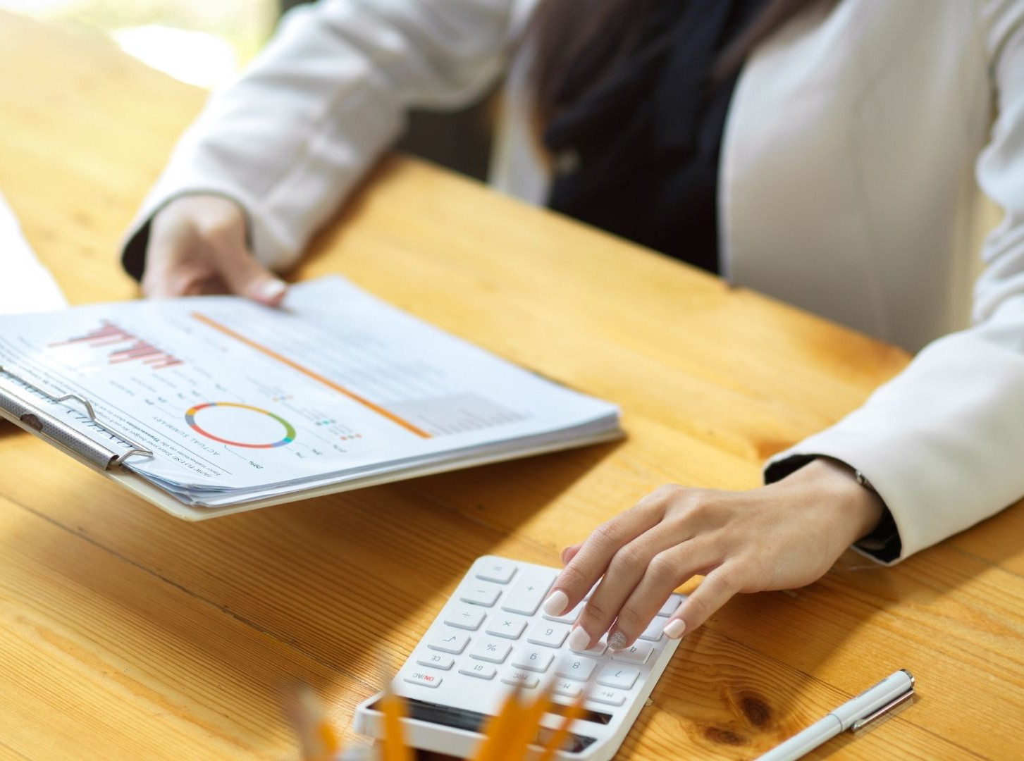 Businesswoman using a calculator beside a financial report and laptop on a sunlit wooden desk, focused on budgeting.