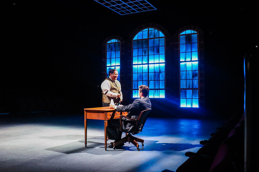 two men engage in serious conversation over a desk in front of ceiling high windows 