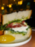 Close-up of a sandwich with pesto, cheese, greens, and cured meat on white bread, with a yellow pepper on a white plate. Dimly lit background. Tiny's Bar Sandwich