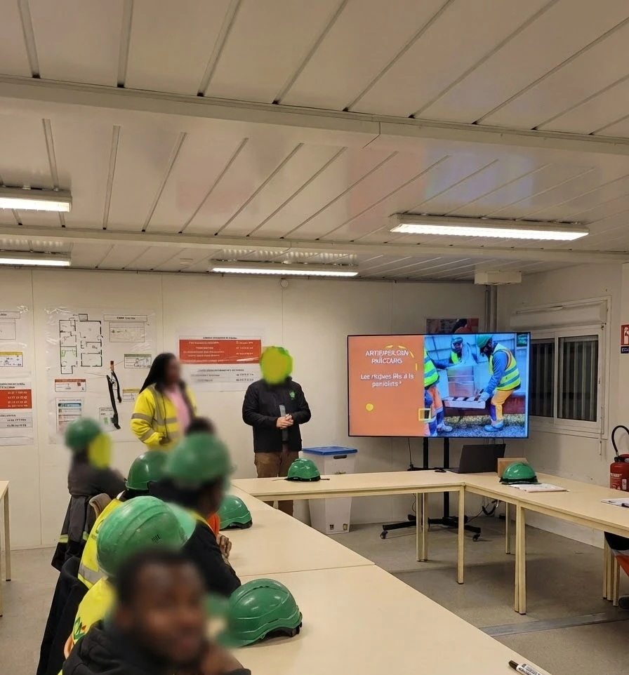 Digital photograph of a safety training session. A presenter addresses a group of people wearing safety helmets and vests in a bright classroom.
