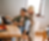 A queer couple shares a joyful moment while working together at home. One partner sits at a laptop, smiling, while the other leans in, engaging warmly in conversation. The scene reflects love, collaboration, and everyday connection