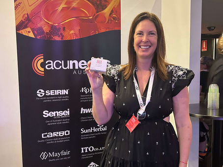 Dr Angela Marshall, Brisbane acupuncturist at Womb to World, in a black dress holding a card in front of an "Acuneeds" banner.