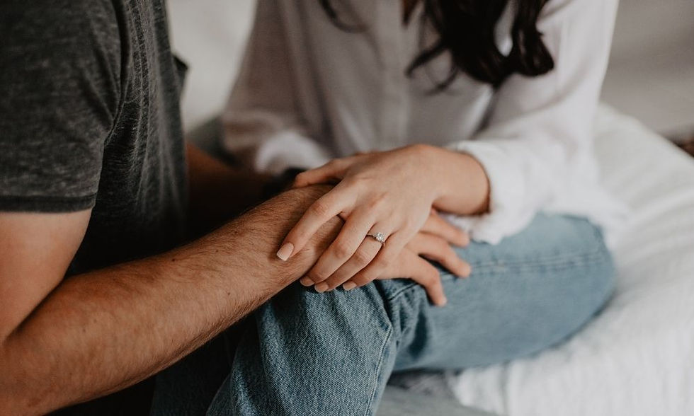 A couple holds hands warmly. The woman wears a ring, and they sit on a white bed. She wears a white shirt, he wears a dark shirt. Possibly anxious about IVF results.