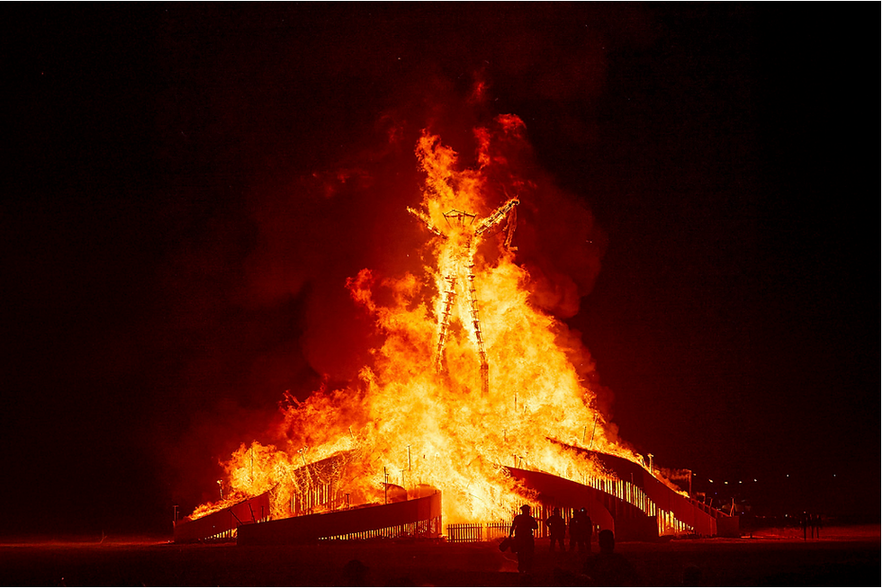 A large wooden figure burns brightly at night, surrounded by silhouetted spectators. Intense orange flames illuminate the dark sky.