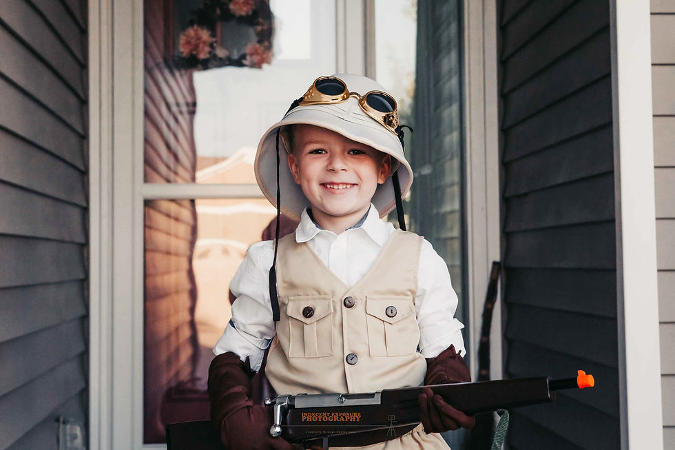 Smiling child in safari outfit with hat and goggles holds toy rifle on a porch. Background features flowers and siding. Casual, playful mood.