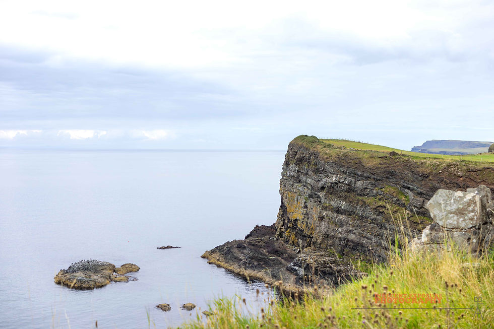 Cliff overlooking calm ocean with cloudy sky. Grass tops the cliff, scattered rocks in the water. Serene, natural setting.