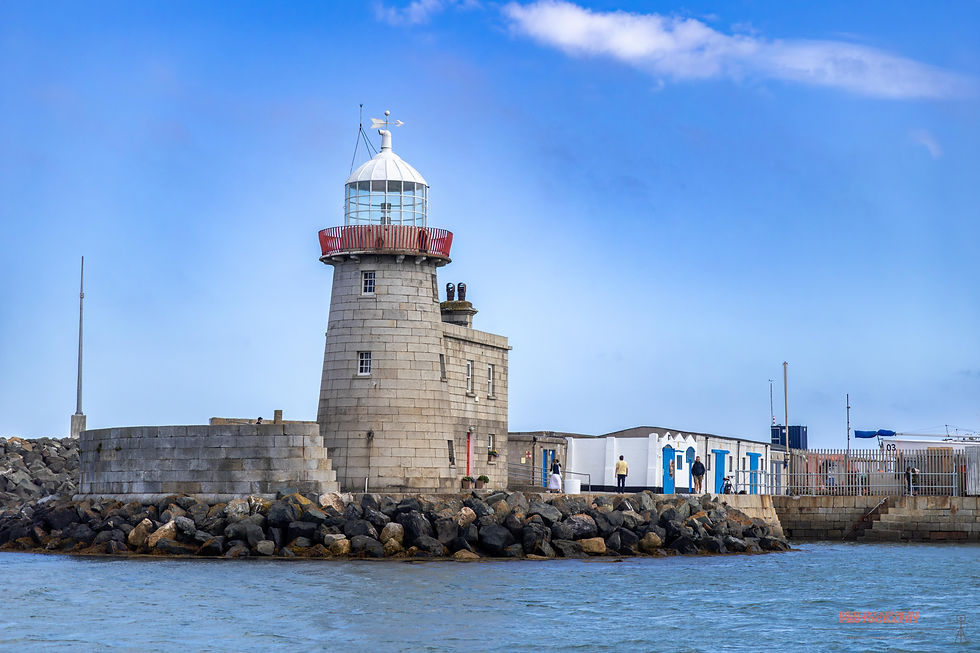 Stone lighthouse with a red railing on a rocky pier, under a clear blue sky. People walk nearby, creating a calm seaside scene.