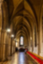 Gothic hallway with vaulted arches, stained glass window, and red-cushioned wooden chairs. People sit and walk, creating a serene atmosphere.