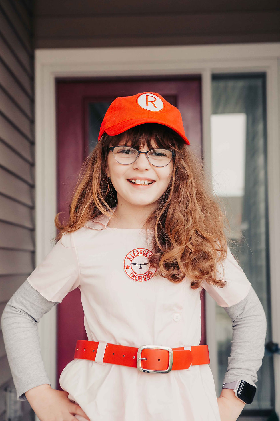 Smiling girl in a red cap and matching belt, wearing a baseball-themed outfit with logo. Stands in front of a maroon door.