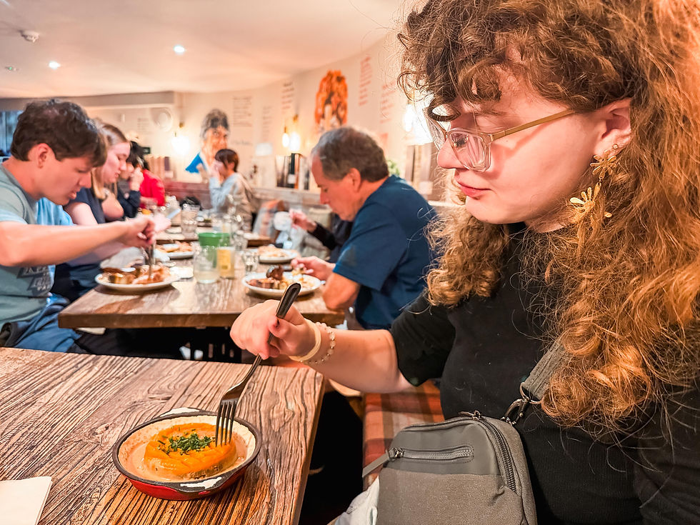 People eating at a wooden table in a cozy restaurant. Woman in glasses focuses on her dish, wearing black top and gold earrings. Warm lighting.
