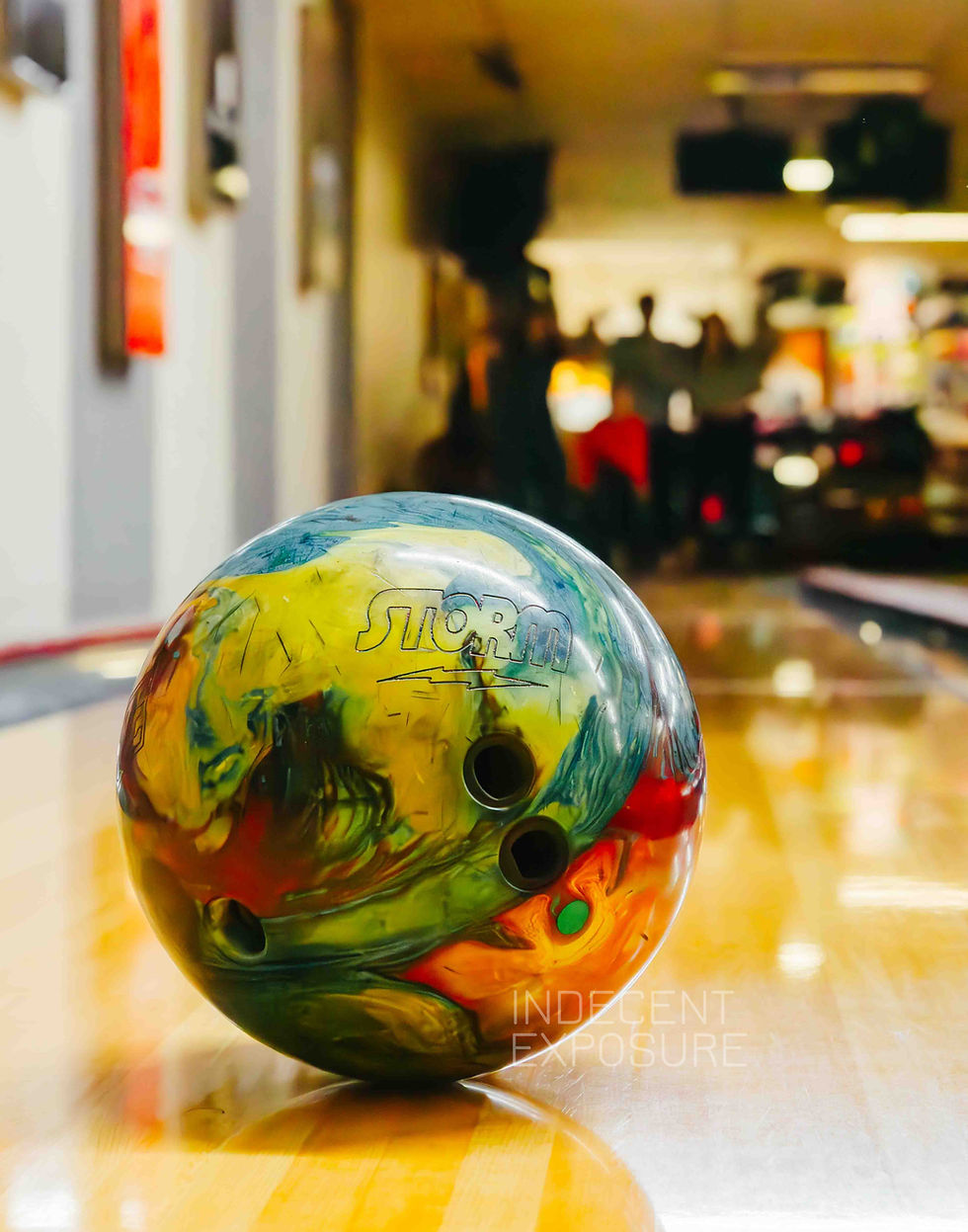 Colorful bowling ball with "Storm" text on a shiny lane, blurred background shows dimly lit bowling alley with people.