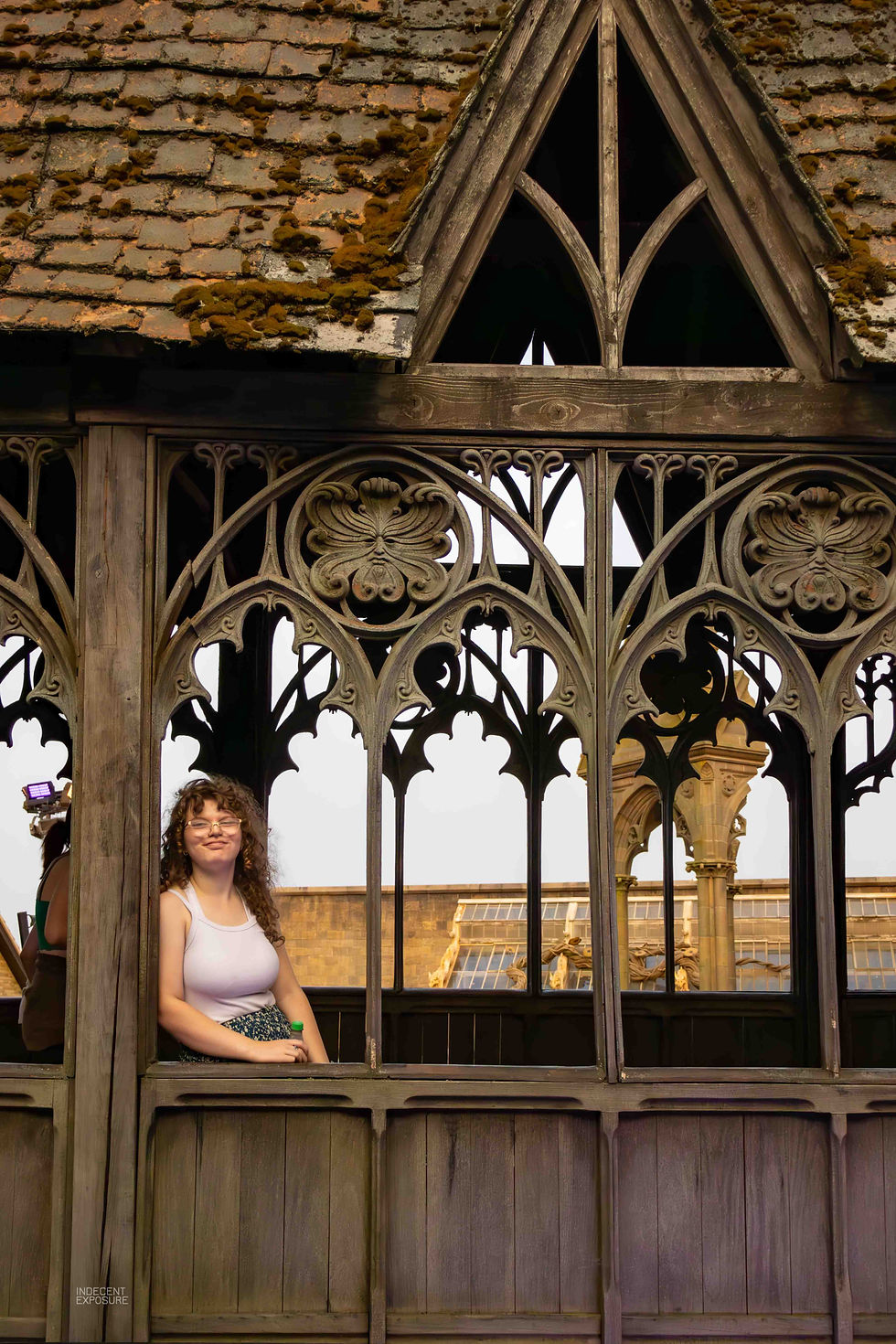 Woman with curly hair smiling through intricate wooden balcony with arched patterns. Background shows a stone building and bright sky.