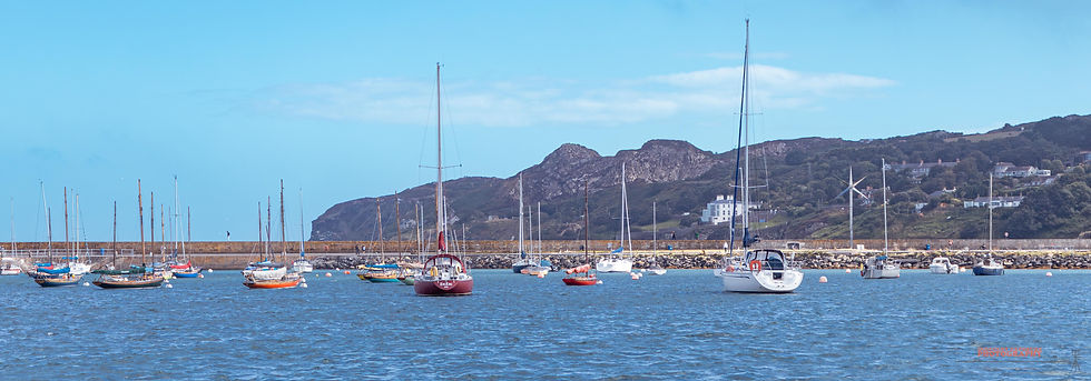 Sailboats float in a calm harbor with mountains in the background under a clear blue sky. A breakwater separates the boats from the shore.