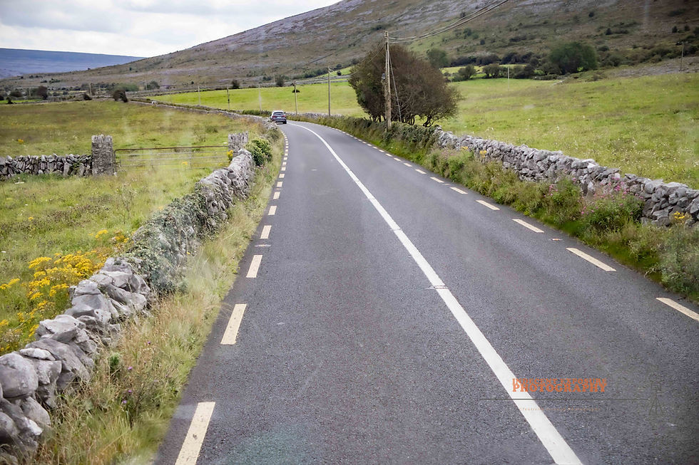 A narrow, winding road flanked by stone walls in a rural, green landscape. A car drives away under a cloudy sky.