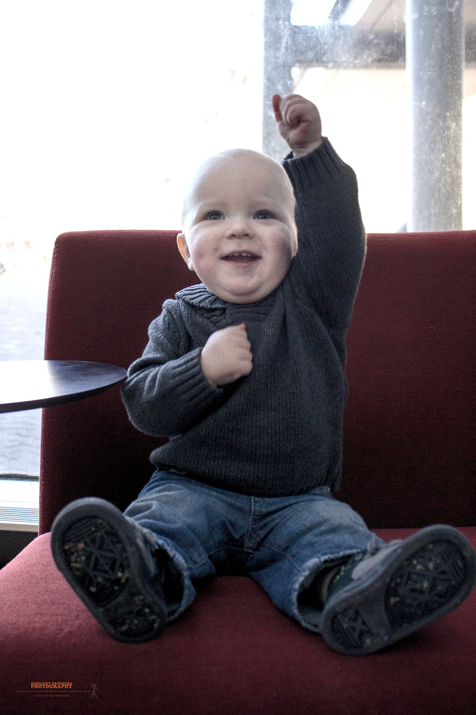 A smiling baby in a gray sweater raises an arm, sitting on a red chair with a sunny outdoor view through a window in the background.