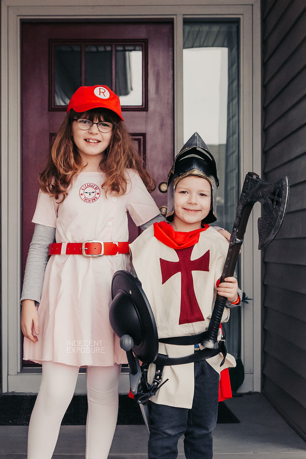 Two kids in costumes stand in front of a red door. One wears sports attire with a red cap, the other a knight outfit with helmet and axe.