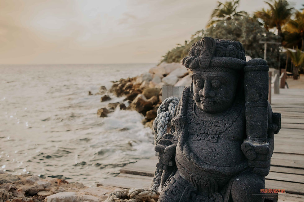 Stone guardian statue on a wooden pier by a calm tropical sea at sunset, with palm trees.