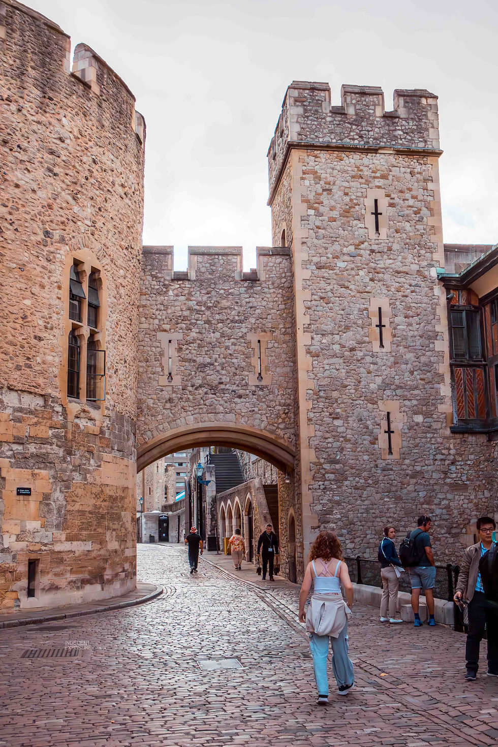 People walk on a cobblestone path through a historic stone archway, with tall stone towers, under a cloudy sky.