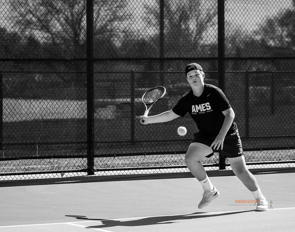 A tennis player in an "AMES" shirt hits a forehand on an outdoor court. The background features a chain-link fence and trees.