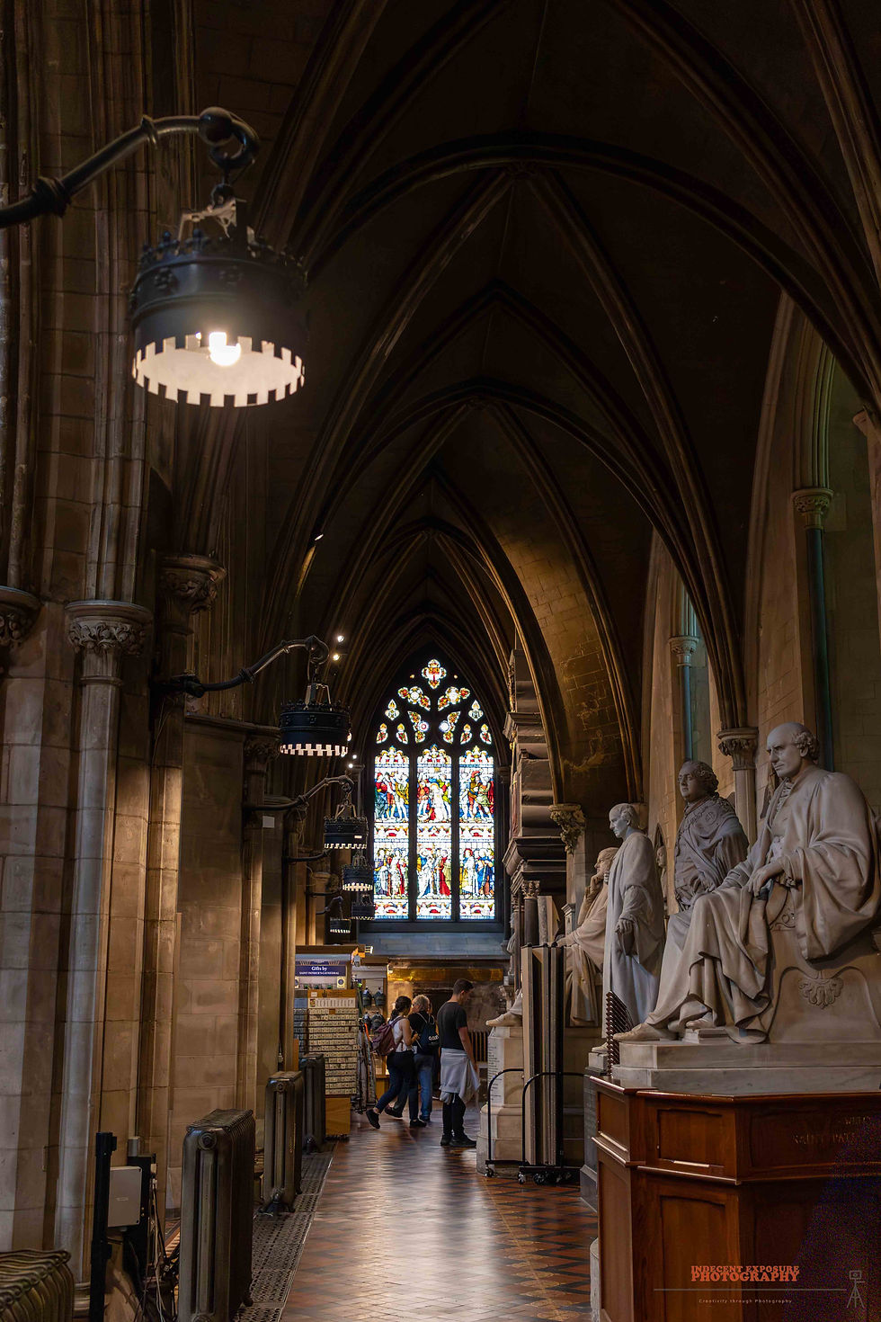 Dimly lit cathedral hall with stained glass window, statues, and arched ceiling. Two people stand beneath lights, adding a serene mood.