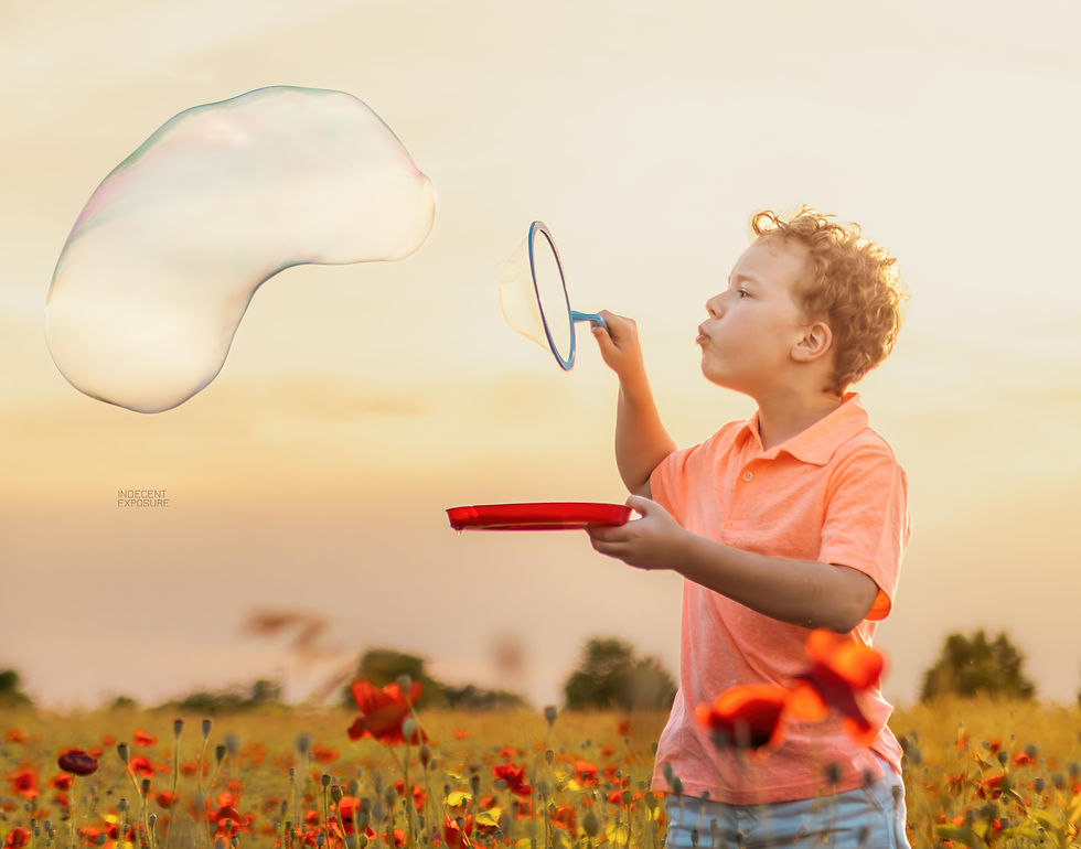 A young boy in a peach shirt blows a bubble in front of a sunset while standing in a field of poppies.