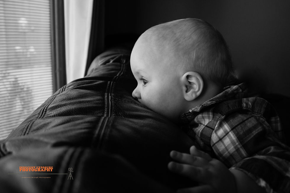 A baby in plaid shirt gazes intently out window, head resting on leather. Soft lighting, serene mood. Blinds and curtain visible.