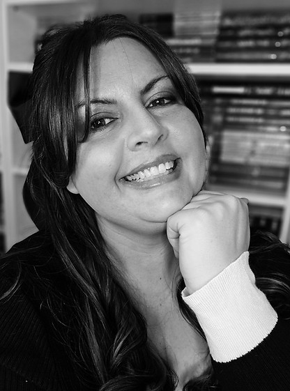 Black and White photo of author Michelle Sauer. Hand beneath her chin with her head tilted to the side, smiling. Long dark hair with a dark bow.