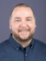 Smiling person with a beard in a blue button-up shirt against a plain gray background.