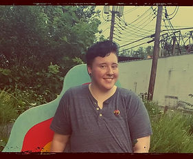 A person in a grey t-shirt stands smiling in front of an abstract backdrop with greenery behind them