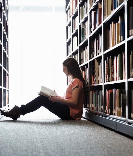 Woman reading on the floor of a library with her back against a book shelf