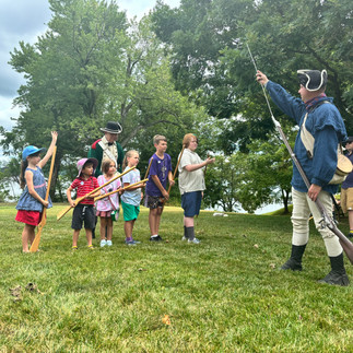 A man in colonial attire teaches a group of kids how to drill with fake bayonets