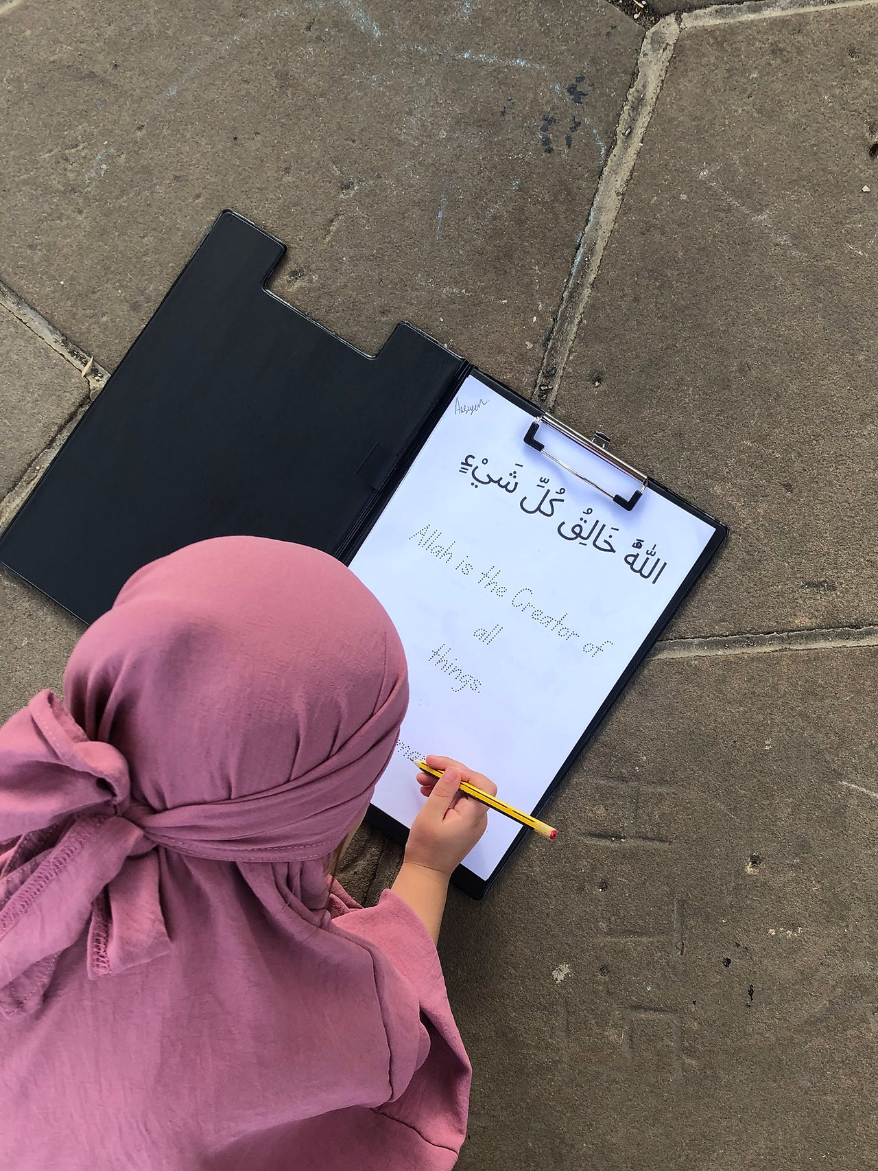 Young Muslim girl wearing a pink hijab completing an Islamic homeschooling worksheet with Arabic text that says ‘Allah is the Creator of all things’ — hands-on learning activity for children.