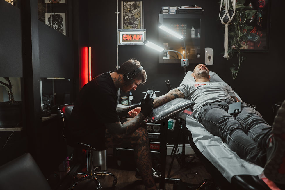 Tattoo artist inks client's arm in a dimly lit studio with red neon "ON AIR" sign. Client lies on a chair, phone on lap. Mood is focused.
