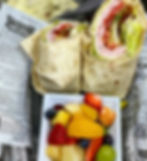 Wrap halves on newspaper wrap next to a fruit bowl with strawberries, oranges, blueberries. Casual and vibrant food presentation.