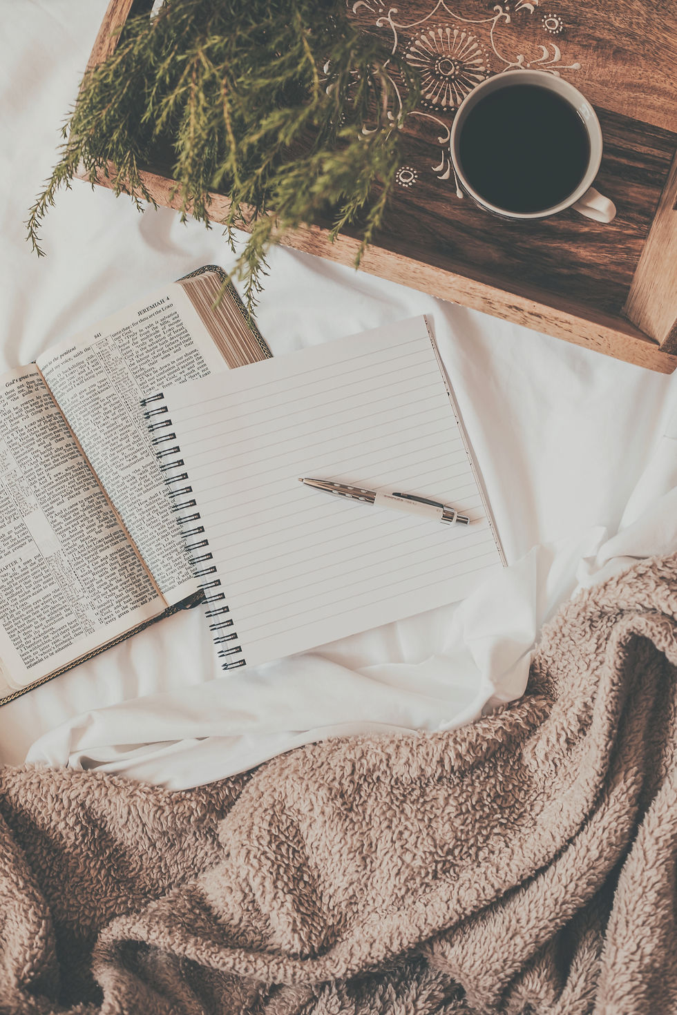 Cozy morning setup with an open book and notepad on soft bedding, complemented by a cup of coffee on a wooden tray and a touch of greenery.