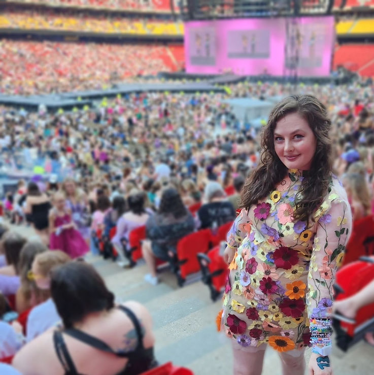 Jennifer stands to the side of a crowded concert venue. Other people are blurred, but Jennifer stands, softly smiling to the right of center wearing a multicolored floral applique dress reminiscent of a Taylor Swift Grammy dress from her folklore era.