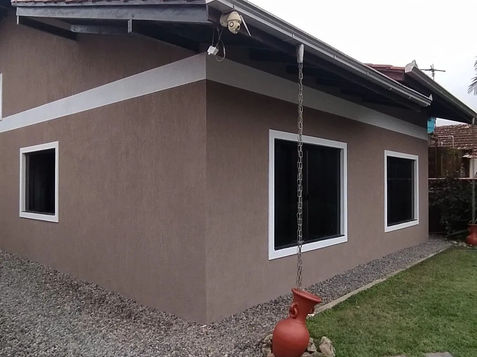 Casa marrom com telhado e janelas brancas, gramado verde e céu ensolarado.