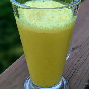 Vaso de batido de leche de cúrcuma sobre una barandilla de madera, con fondo verde desenfocado. El batido tiene espuma en la parte superior.