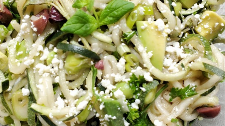 Zucchini noodles with avocado, feta, and herbs in a clear bowl. Fresh greens and white cheese are sprinkled, giving a vibrant look.