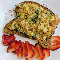 Toast topped with a creamy salad mix, featuring diced tomatoes and greens, on a white plate with sliced strawberries on the side.