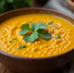 Orange lentil soup in a brown bowl garnished with fresh parsley. Wooden table background with scattered lentils and herbs. Cozy vibe.
