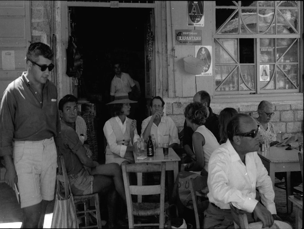 People - including Charmian Clift and George Johnston - relax at an outdoor café, some seated at tables, others standing. Posters on stone walls, vintage vibe, casual mood.