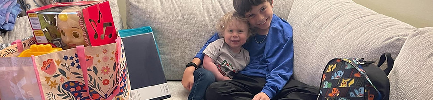 two boys sit on a couch smiling holding eachother next to a bag of toys