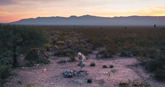 A man standing by a fire pit in the middle of the dessert during sunrise.