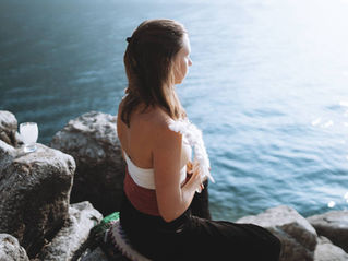 A woman sits pensively on rocks near water.