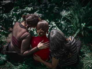 Three woman with their backs to us, wrap their arms around each other, looking out onto a forest of plants and ferns.