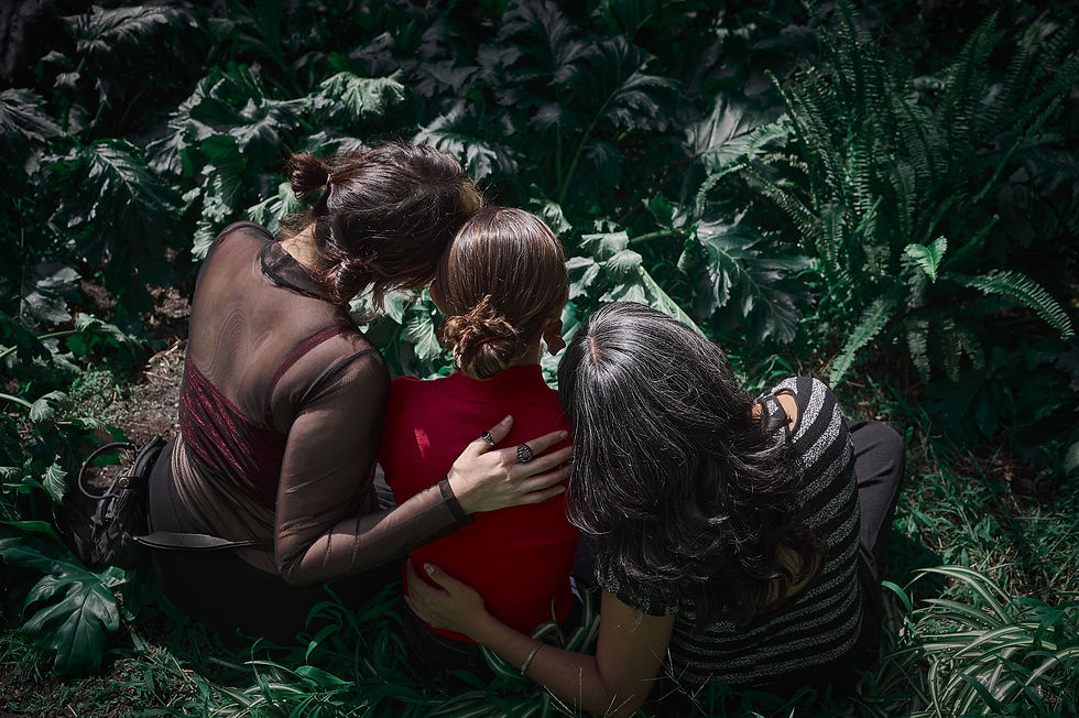 Three woman with their backs to us, wrap their arms around each other, looking out onto a forest of plants and ferns.
